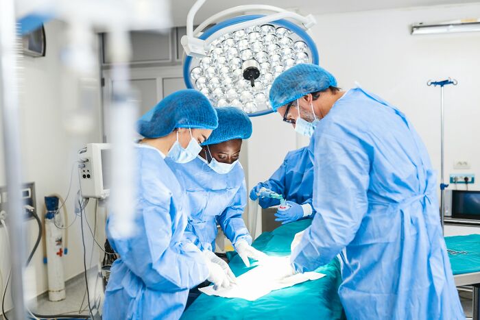 Medical team in surgical gowns and masks performing a procedure under bright operating room lights inside a hospital.