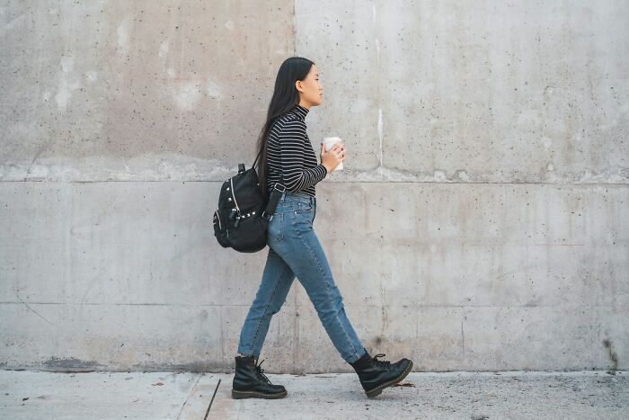 Young woman walking with coffee cup and backpack against plain wall, illustrating things normal in America but offensive elsewhere