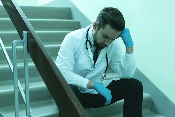 Male doctor wearing gloves and a stethoscope sitting on stairs, appearing stressed about unethical actions with a patient.