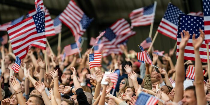 Crowd waving American flags enthusiastically at an outdoor event, highlighting things normal in America but offensive elsewhere.