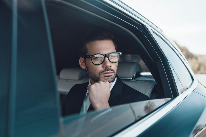 Young man with glasses in a suit looking thoughtfully out the car window reflecting on adult goals and life choices.
