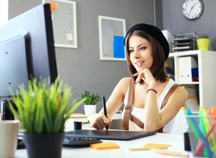 Young woman wearing a black hat, using a digital pen and tablet while smiling at her desktop in a creative office space.