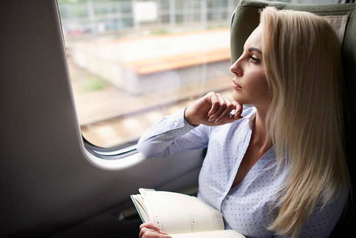 Woman with long blonde hair sitting by a window, holding a book and looking thoughtfully outside while coping with triggers.