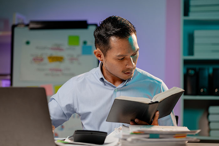Young gifted man focused on reading a book at desk, surrounded by study materials and a laptop in a home office.