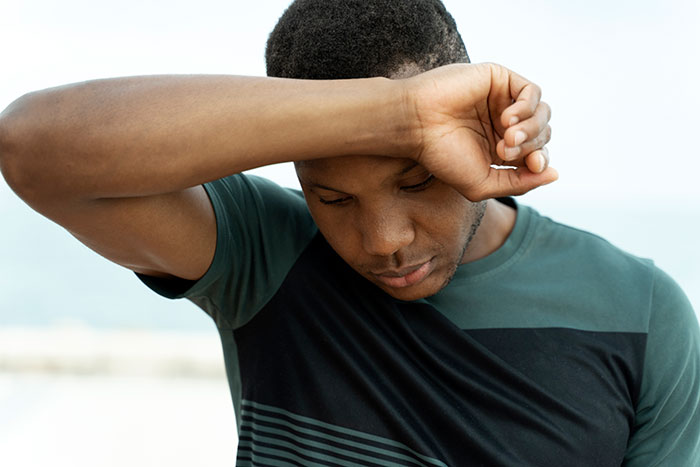 Young gifted man in casual wear wiping forehead, reflecting on potential and achievements outdoors on a bright day.