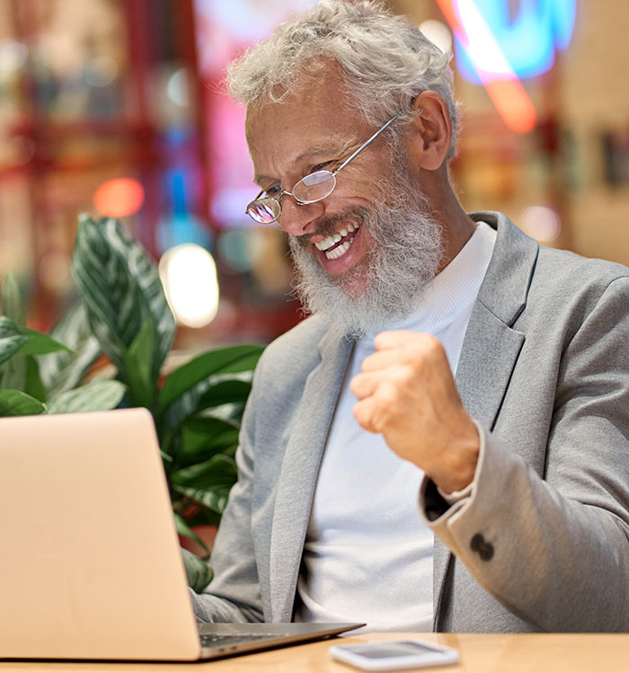 Older man with glasses, smiling and celebrating success while working on laptop, illustrating gifted kids' potential.