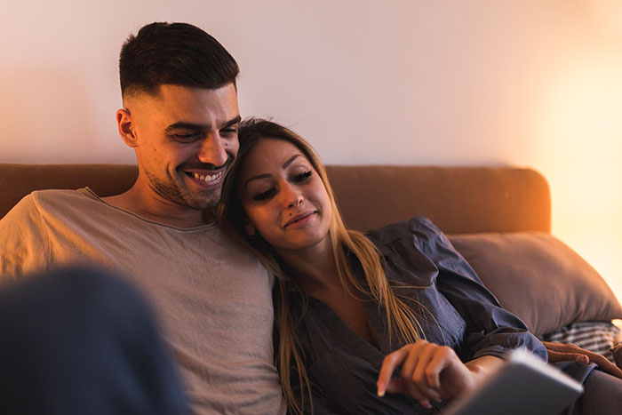 Couple on a couch with the man showing lovey-dovey affection while the tired girlfriend looks fed up with morning kisses.