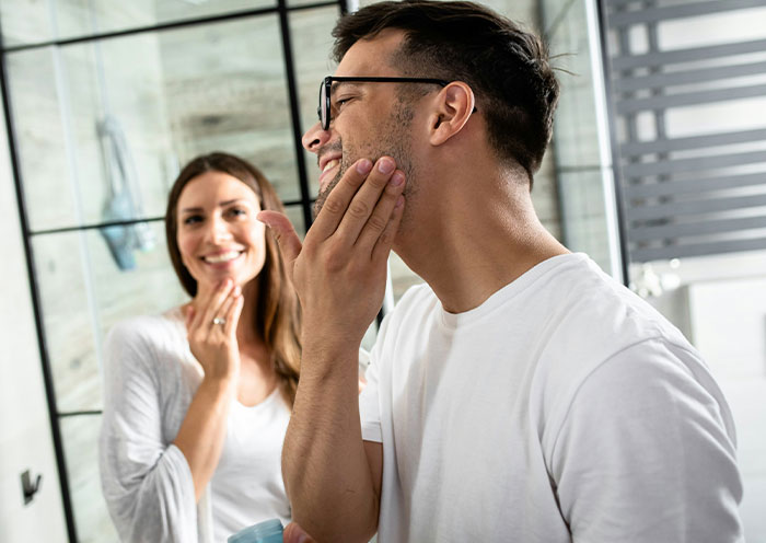 Man shaving in bathroom mirror while woman watches, illustrating woman demands swimmer boyfriend look like her friend’s partner.