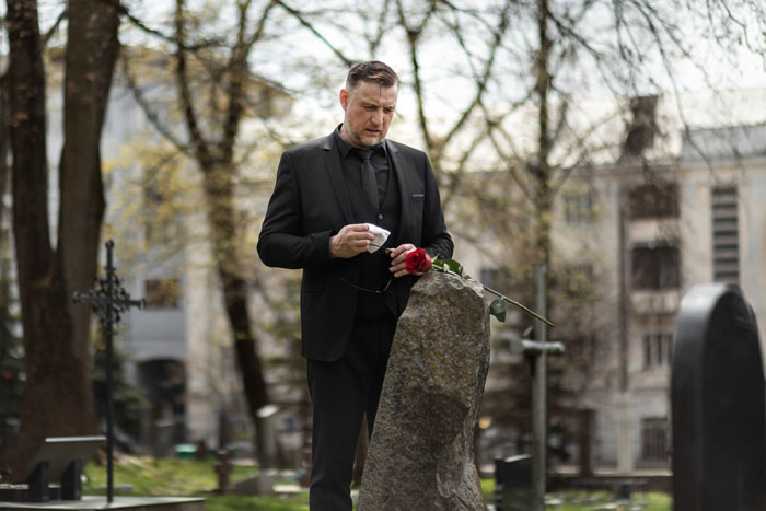 Man grieving at tombstone in cemetery holding rose, reflecting on loss and emotional demands during mourning. Man grieving at tombstone in cemetery holding rose, reflecting on loss and emotional demands during mourning.