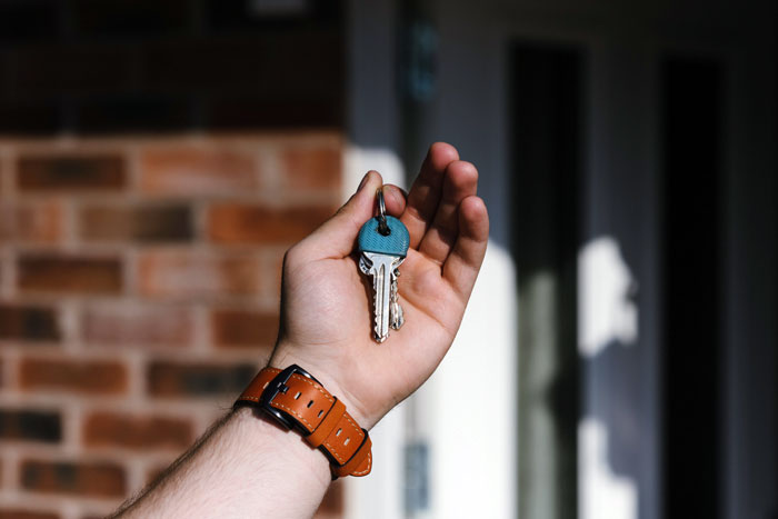 Hand holding house keys with a blue tag in front of a brick wall, symbolizing hidden key and home entry concerns.