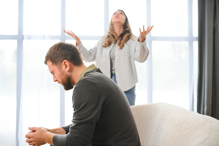 Man upset sitting on couch with woman in background expressing frustration about family and immature behavior Man upset sitting on couch with woman in background expressing frustration about family and immature behavior