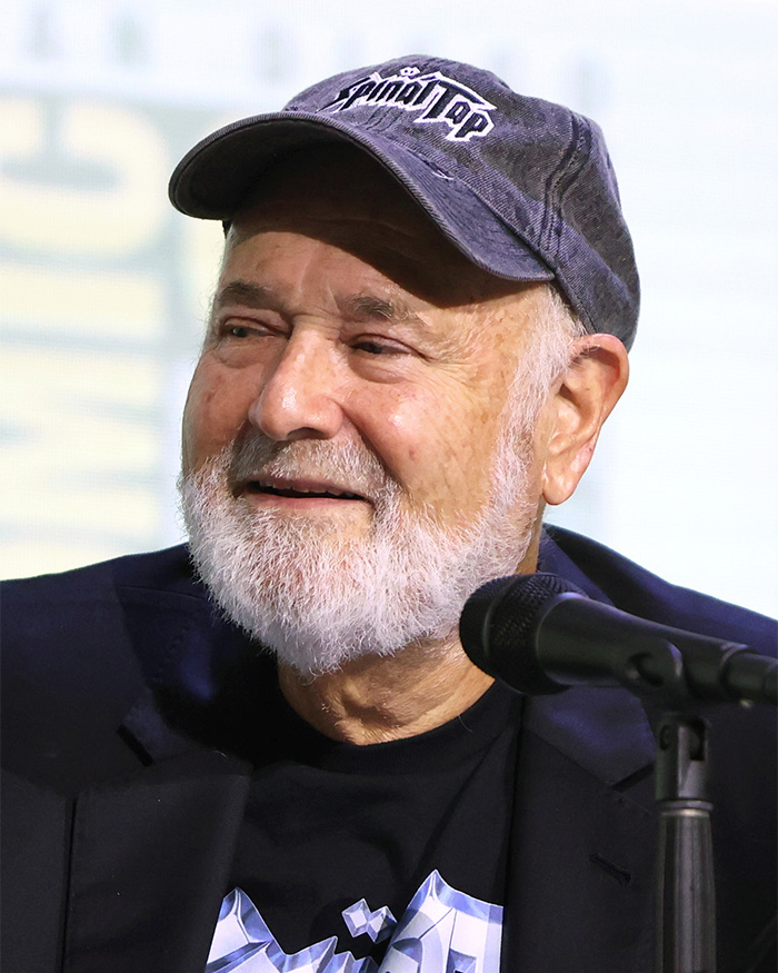 Elderly man with white beard wearing a cap speaking at a microphone during a Golden Globes event panel.