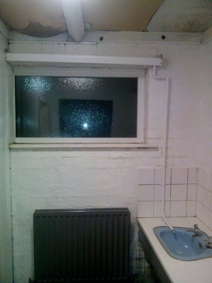 Poor DIY plumbing with a pipe awkwardly installed from ceiling to a sink in a small, dimly lit room with a radiator below a window.