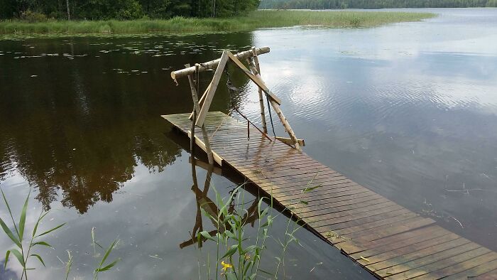 Unstable wooden dock failing at DIY with poorly constructed handrails over calm lake water surrounded by greenery.