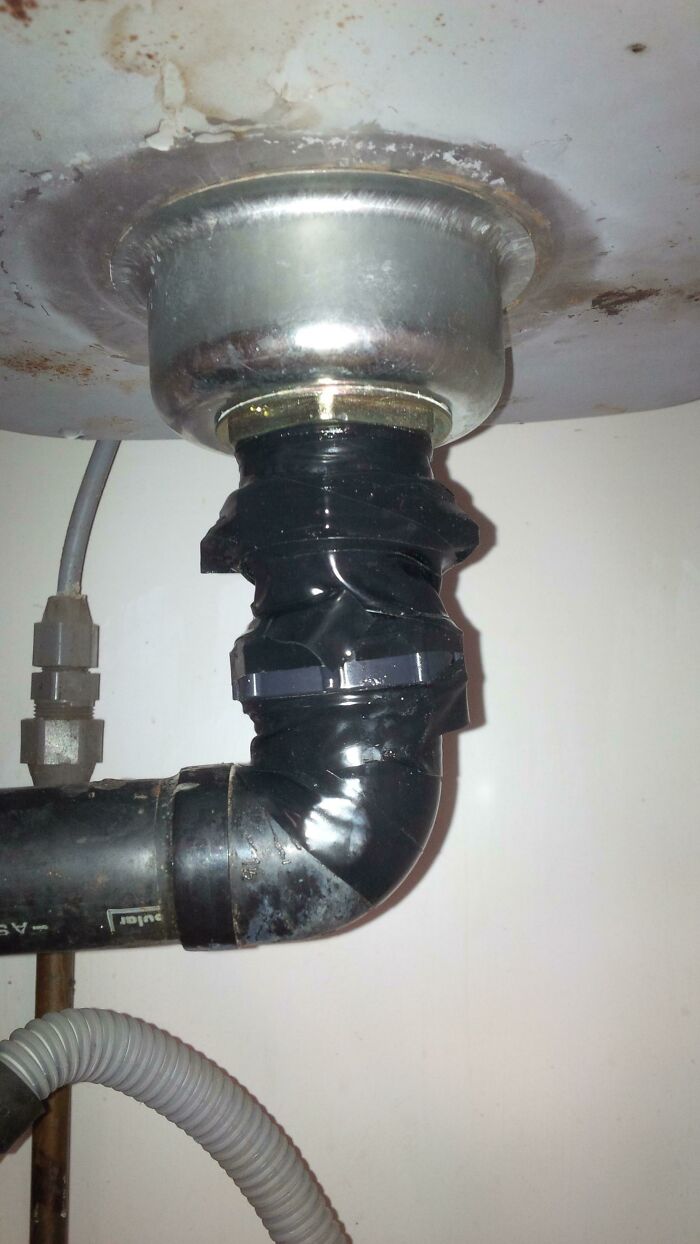 Improvised DIY plumbing repair under a sink using black tape to fix leaking pipes unsuccessfully.