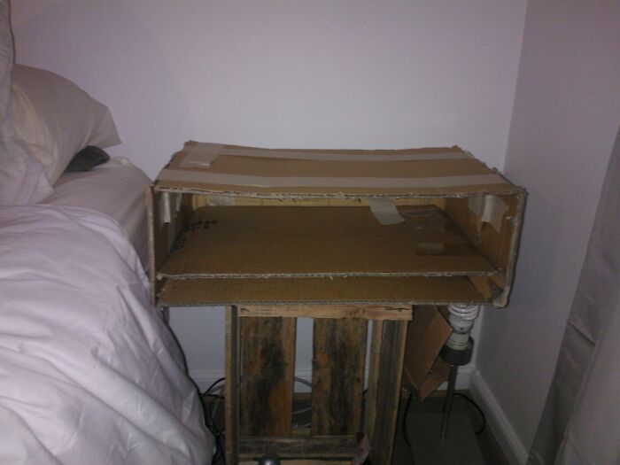 Homemade cardboard DIY shelf placed next to bed, showing a poorly executed and unstable construction project.