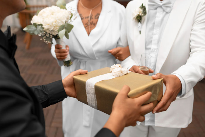 Person in black shirt handing a gold wrapped wedding gift to a bride and groom dressed in white attire.