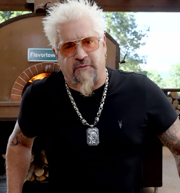 Guy Fieri wearing sunglasses and a chain necklace, standing in front of a brick pizza oven in Flavortown.