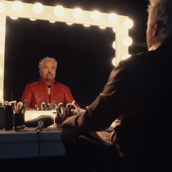 Guy Fieri sitting at a vanity mirror with glowing lights, reflecting on his 58th birthday glow-up and hair transformation.