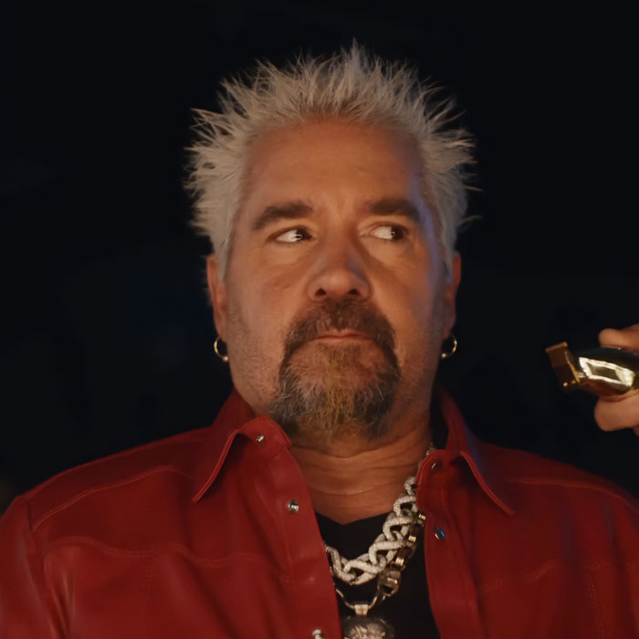 Guy Fieri with spiky blonde hair, wearing a red leather jacket and chain necklace in a dimly lit setting. Guy Fieri with spiky blonde hair, wearing a red leather jacket and chain necklace in a dimly lit setting.
