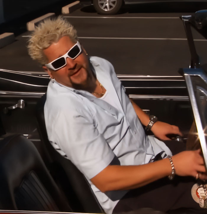 Guy Fieri sitting in a car wearing white sunglasses and a light blue shirt, showing his spiky blonde hair style. Guy Fieri sitting in a car wearing white sunglasses and a light blue shirt, showing his spiky blonde hair style.