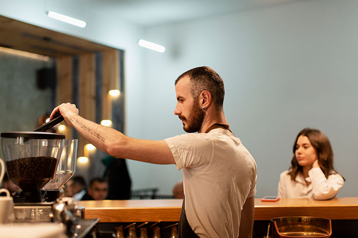 Barista preparing coffee behind counter while a woman waits, illustrating coffee line patience and morning routine.