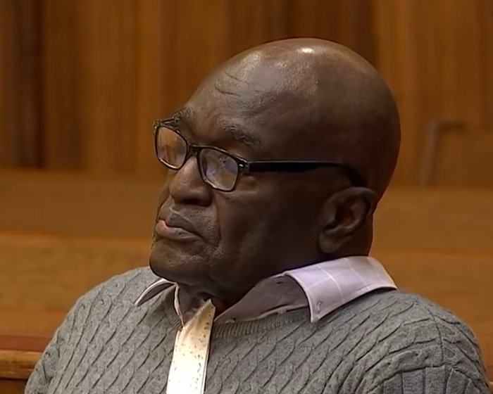 Elderly man wearing glasses and a gray sweater, listening intently in a wood-paneled courtroom setting about Alligator Alley case.