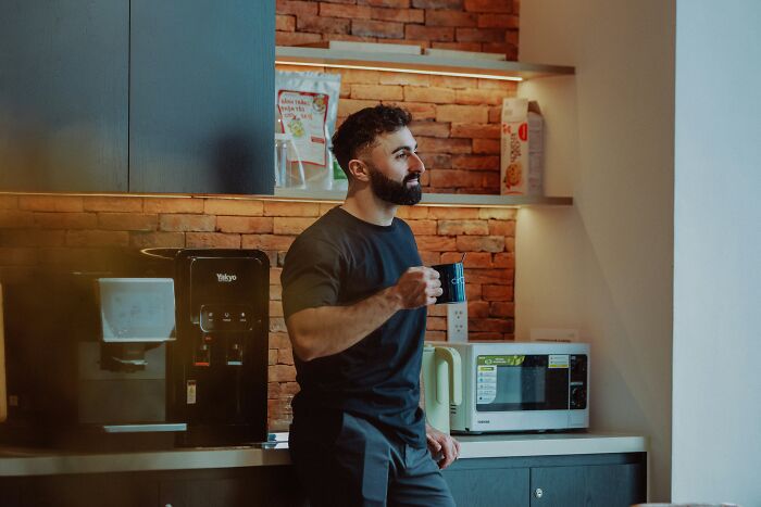Man in a black shirt holding coffee cup in office kitchen sharing stories about coworkers acting unhinged and unprofessional.
