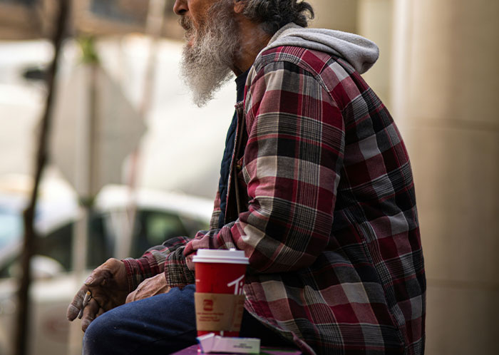Homeless man with a beard in a plaid shirt sitting outdoors with a coffee cup, illustrating homeless people experiences.