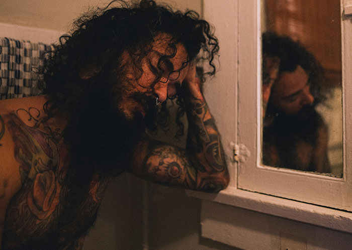 Homeless man with tattoos and curly hair reflecting sadly in a mirror, illustrating struggles of homeless people experiences.