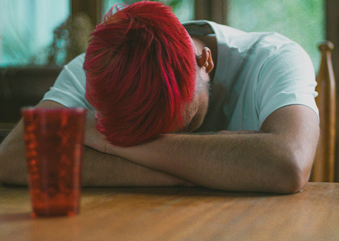 Young man with red hair resting head on folded arms on a wooden table, reflecting the struggles of homeless people.