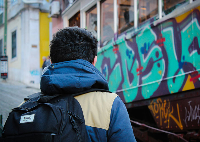Person with backpack and hooded jacket walking past a graffiti-covered wall, illustrating homeless people experiences.