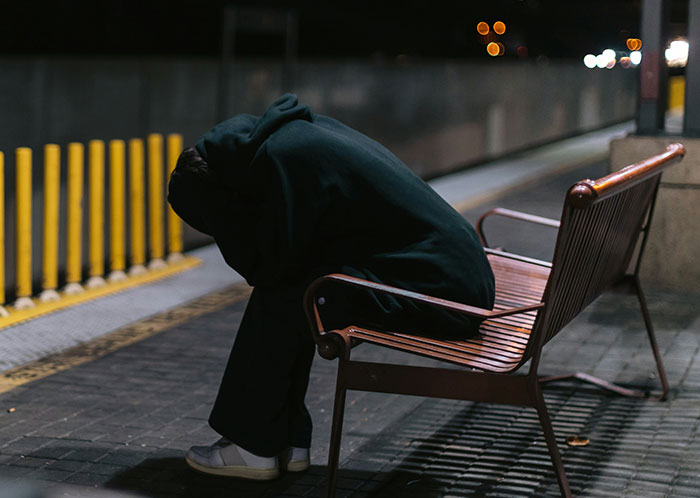Person experiencing homelessness sitting on a bench at night, reflecting the harsh realities of homeless people’s experiences.