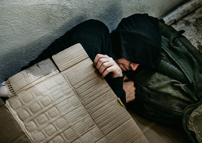 Homeless person in a black hoodie curled up on the ground with bags and cardboard, highlighting harsh homeless experiences.