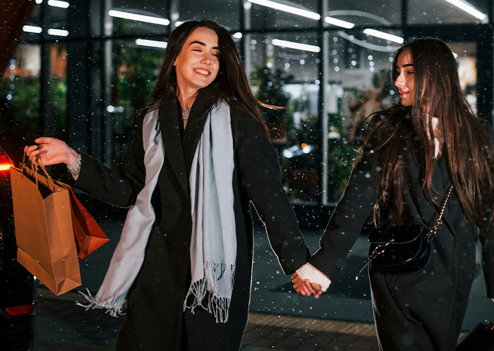 Two women smiling and holding hands outside at night in winter coats, unrelated to homeless people experiences.