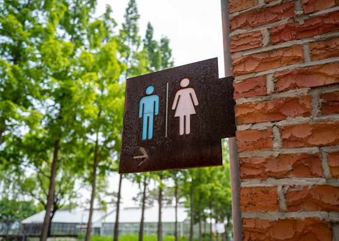 Restroom sign on a brick wall outdoors with trees in the background, highlighting challenges faced by homeless people.