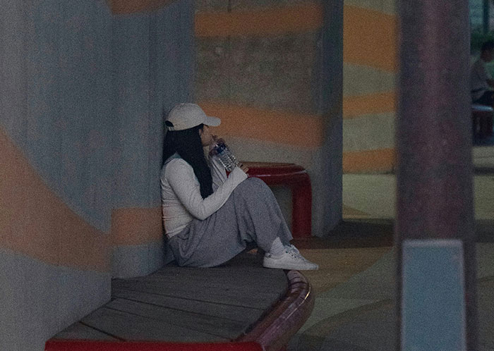 Young homeless person sitting alone on bench at night, wearing casual clothes and drinking water, reflecting harsh experiences.
