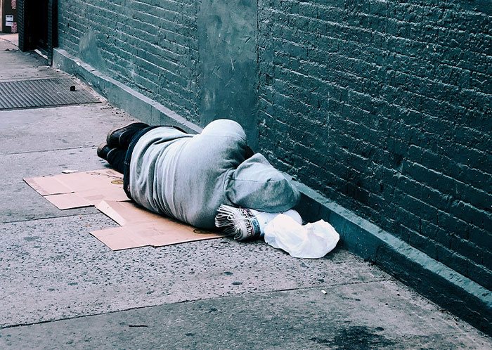 Homeless person lying on cardboard on a city sidewalk against a brick wall, highlighting homeless people experiences
