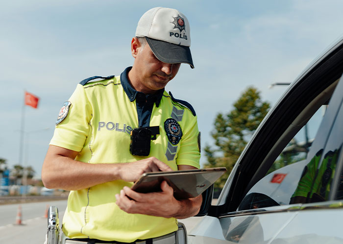 Police officer in uniform writing a report on a tablet during a traffic stop near a white car outdoors.