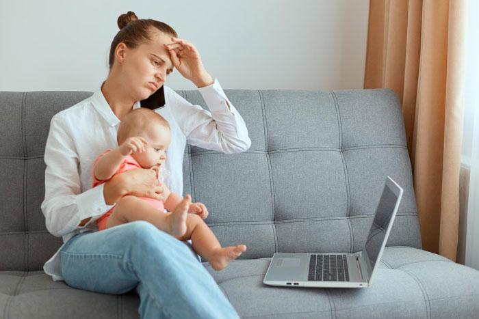 Stressed woman holding baby on couch, looking upset while working on laptop, illustrating child-loathing and nanny struggles. Stressed woman holding baby on couch, looking upset while working on laptop, illustrating child-loathing and nanny struggles.