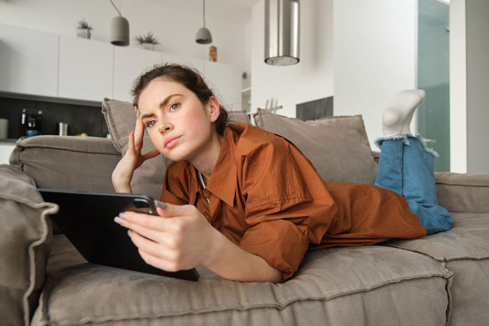 Young woman lying on couch, looking thoughtful and concerned while holding a tablet about inherited home issues.