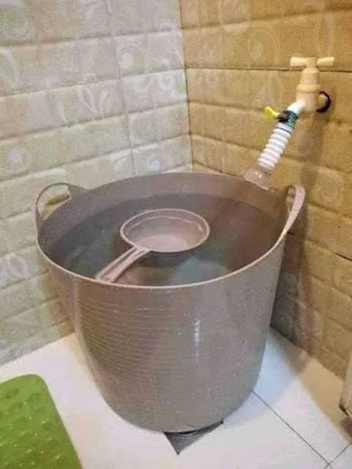 Large plastic bucket filled with water and a strainer, connected to a wall faucet, unique household item in some countries.
