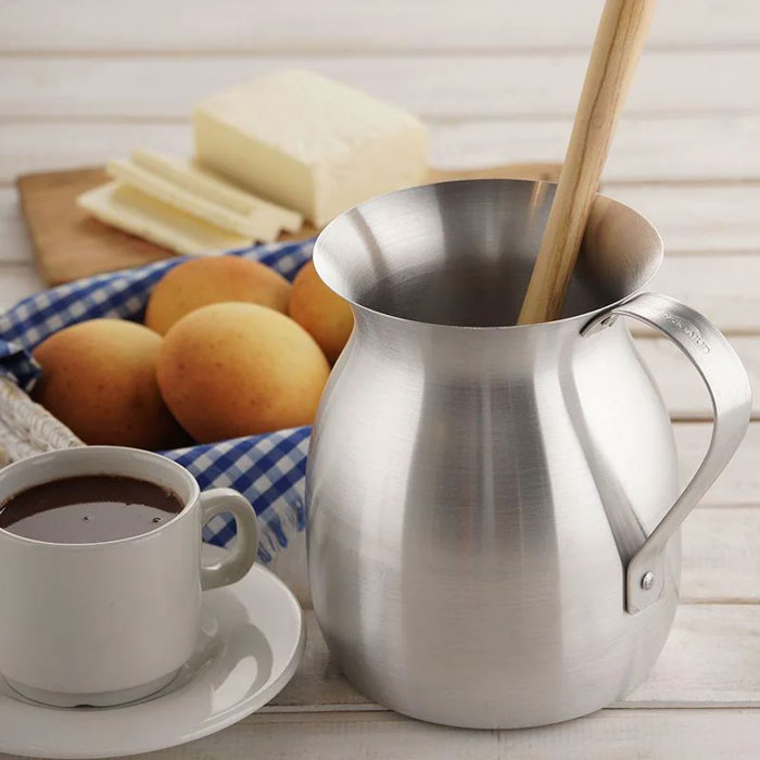 Metal milk frother with wooden stirrer, cup of hot chocolate, bread rolls, and butter on a wooden table showcasing unique household items.