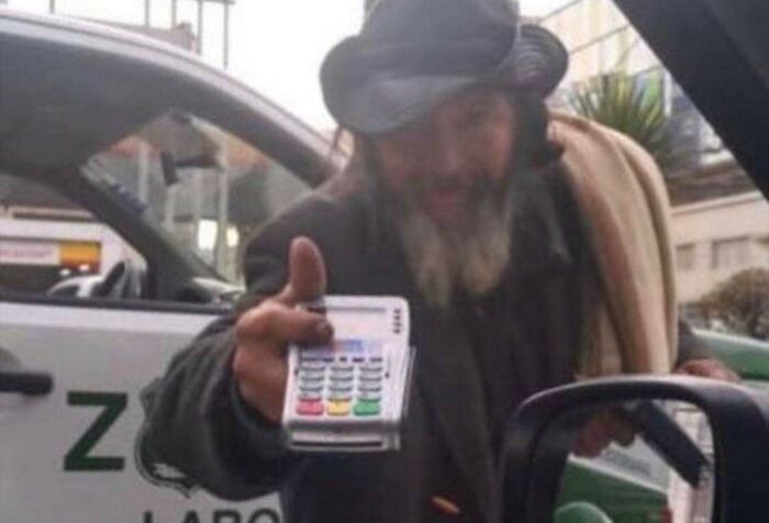 Man with a beard and hat holding a card reader outside a car, illustrating concepts of modern dystopia and strange normality.