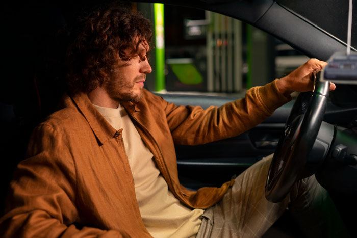 Man with curly hair wearing a brown jacket sitting in a car, portraying a casual boyfriend in a complicated situationship.