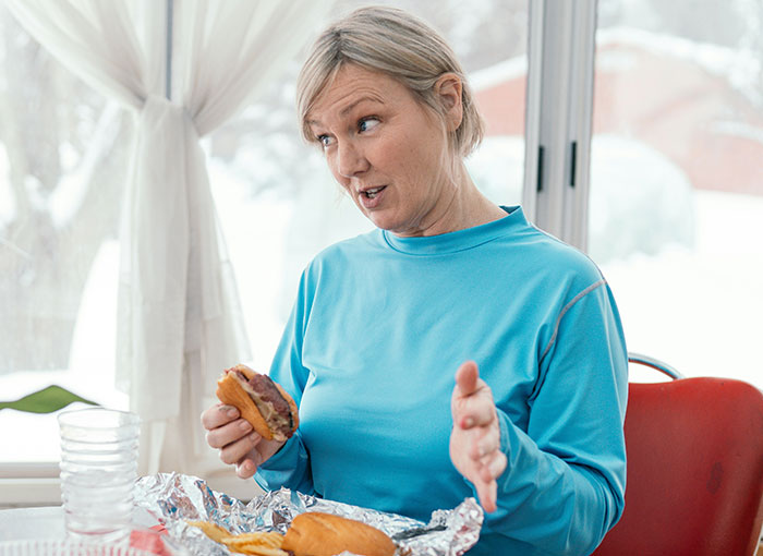 Woman in a blue shirt eating a sandwich while discussing a fun fact with a child and his dad nearby. Woman in a blue shirt eating a sandwich while discussing a fun fact with a child and his dad nearby.