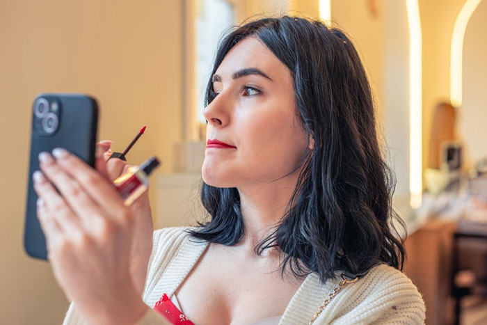 Young woman confidently applying lipstick while looking in her phone mirror, expressing her autonomy despite toxic behavior.