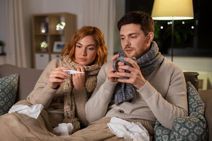 Wife looking at thermometer while husband with scarf holds mug, showing fed up feelings toward insufferable sick husband.