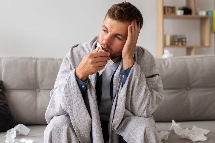 Sick husband wrapped in blanket looking insufferable on couch surrounded by tissues, reflecting fed up wife’s frustration. Sick husband wrapped in blanket looking insufferable on couch surrounded by tissues, reflecting fed up wife’s frustration.