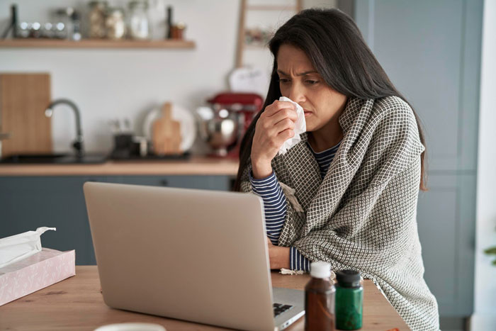 Woman wrapped in a blanket looking frustrated while using laptop, illustrating fed up wife dealing with insufferable sick husband.